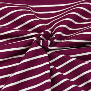 Può includere: Primo piano di un tessuto con un motivo ripetuto di strisce bianche su sfondo magenta scuro. Il tessuto è raccolto e piegato, creando un effetto strutturato. Le strisce corrono in diagonale.