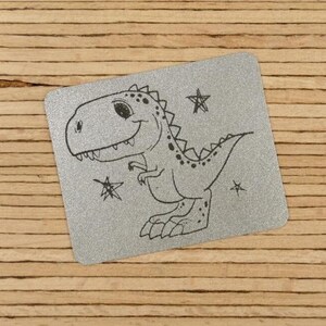 Könnte beinhalten: Ein rechteckiger, silberfarbener Metallanhänger mit einer Cartoon-Dinosaurier-Illustration und Sternen. Der Dinosaurier lächelt und hat ein gepunktetes Muster auf dem Rücken. Der Anhänger befindet sich auf einem Holzgrund.