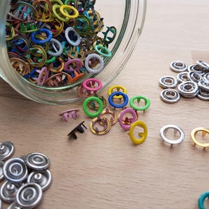 Könnte beinhalten: Eine Sammlung bunter Metallösen und Druckknöpfe, darunter silberne, goldene und verschiedene Farbtöne von Blau, Grün, Rosa und Gelb. Einige Ösen liegen auf einer Holzoberfläche verstreut, andere in einem Glas. Diese Verschlüsse sind zum Basteln und Nähen.