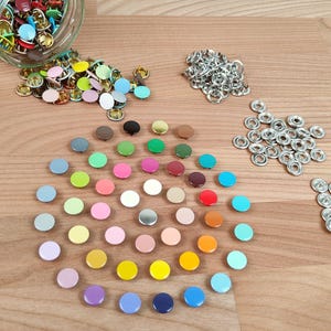 Könnte beinhalten: Eine Sammlung bunter Metall-Druckknöpfe und Ösen in verschiedenen Farben, darunter Blau, Grün, Rosa und Gelb. Einige sind auf einer Holzoberfläche verstreut, andere in einem Glas. Die Verschlüsse werden für Kleidung und Bastelprojekte verwendet.