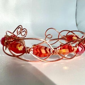 May include: A handcrafted copper wire bracelet featuring red and orange faceted beads. The open cuff design has decorative swirls and woven sections, creating an intricate and artistic look. The bracelet is designed to be worn on the wrist.