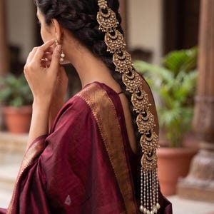 May include: A gold-toned hair accessory featuring a series of crescent-shaped ornaments adorned with small white stones, arranged vertically along a braided hairstyle and finished with a dangling fringe of white pearl-like beads at the bottom.