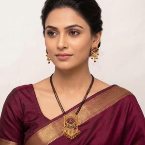 Puede incluir: Conjunto de collar y pendientes dorados. El collar presenta un colgante rectangular con una piedra roja central, rodeada de piedras verdes y rojas más pequeñas. Los pendientes tienen un diseño similar, con detalles rojos y verdes.