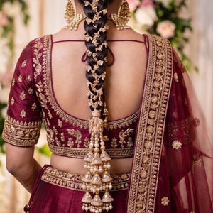 May include: A deep maroon blouse and skirt set featuring intricate gold embroidery. The blouse has a scoop back and short sleeves. A long, dark braided hairstyle is adorned with gold hair accessories and a multi-tiered pearl and gold tassel ornament.