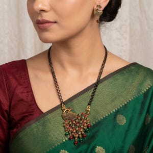 May include: A gold-toned necklace featuring a peacock-shaped pendant with red and green stones and dangling beads, paired with matching peacock stud earrings. The necklace has a double strand of small black beads and a textured metal finish.