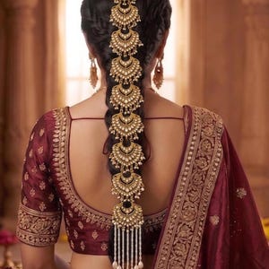 May include: A long gold hair accessory featuring a series of stacked, crescent-shaped filigree ornaments adorned with small white beads and pearls, worn down a braided hairstyle, paired with matching gold drop earrings and a deep maroon blouse.