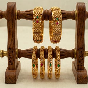 Op de afbeelding: Een houten sieradenstandaard houdt zes gouden armbanden vast. De armbanden zijn versierd met kleine witte kralen en kleurrijke accenten van rode, groene en heldere stenen. De standaard is gemaakt van donker hout met messing beslag.