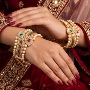 May include: Gold-toned bangles with intricate designs, featuring pearls, red, green, and clear gemstones. The bangles are adorned with detailed patterns and are worn on a person's wrists. The background is a rich red color.