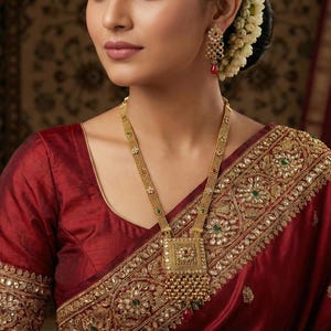 May include: A woman wearing a gold necklace with a square pendant and matching earrings. The necklace and earrings are decorated with small gemstones. She is wearing a red blouse and a red sari with gold embroidery.