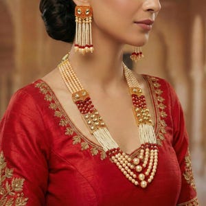 May include: A woman wearing a red blouse and ornate gold jewelry. The jewelry includes a multi-strand pearl necklace with gold and red accents, and matching earrings. The earrings and necklace feature intricate designs and are likely made of precious metals and gemstones.