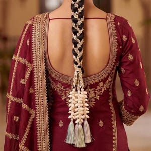May include: A deep maroon long-sleeved garment featuring intricate gold embroidery along the neckline, sleeves, and edges. A dark braided hairstyle is adorned with a gold leaf-patterned hair accessory, white shell-like beads, and tassels.
