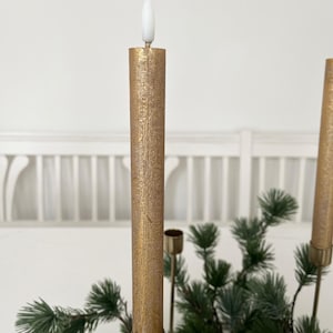 Könnte beinhalten: Goldfarbene, batteriebetriebene Kerze mit einer weißen, flammenförmigen LED-Spitze. Die Kerze hat eine strukturierte Oberfläche und sitzt in einem goldfarbenen Halter. Die Kerze ist Teil einer Weihnachtsdekoration mit grünen Tannenzweigen.