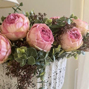 Könnte beinhalten: Nahaufnahme eines Blumenarrangements mit großen, rosa Rosen mit gelben Rändern, umgeben von grünem und braunem Laub. Die Blumen befinden sich in einem strukturierten, cremefarbenen, gerippten Pflanzgefäß, das eine weiche, natürliche Ästhetik erzeugt.