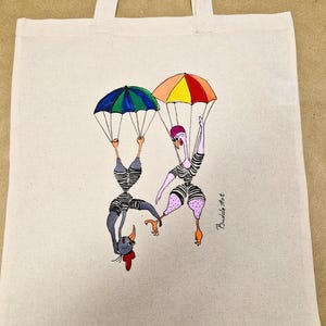 Könnte beinhalten: Beige Canvas-Tragetasche mit einer skurrilen Illustration von zwei Figuren mit vogelartigen Merkmalen, die jeweils an einem bunten Fallschirm hängen. Die Figuren tragen gestreifte Outfits. Die Signatur des Künstlers, Budde Art, ist sichtbar.