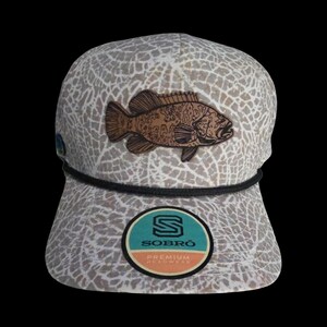 May include: A beige and white patterned baseball cap with a brown fish patch. The cap has a black rope detail and a circular patch with the text "SOBRO PREMIUM HEADWEAR".