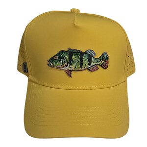 May include: Yellow baseball cap featuring a detailed illustration of a fish. The fish is primarily green and black with red accents. The cap has a curved brim and small perforations on the sides for ventilation. A small logo is visible on the side.