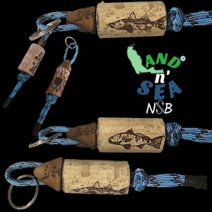 May include: Keychains featuring cork and wood designs with fish illustrations. The keychains have blue and black rope accents and metal key rings. The image also includes the text "AND n' SEA NSB".