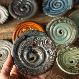 Könnte beinhalten: Eine Sammlung handgefertigter Keramikuntersetzer mit einem Spiraldesign. Die Untersetzer sind in verschiedenen Farben erhältlich, darunter Blau, Grün, Grau und Orange. Die Untersetzer sind rund und haben ein erhabenes Spiralmuster in der Mitte.