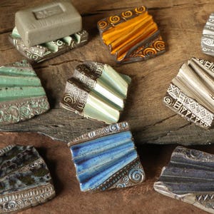 Könnte beinhalten: Eine Sammlung handgefertigter Keramik-Seifenschalen in verschiedenen Farben, darunter Grün, Blau und Orange. Jede Schale hat ein einzigartiges strukturiertes Design und dekorative Kanten, die eine rustikale, künstlerische Ästhetik zeigen. Einige Schalen enthalten Seife.