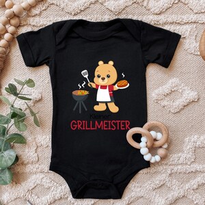 Könnte beinhalten: Schwarzer Baby Strampler mit einem Cartoon-Bär, der ein Hotdog grillt. Der Text auf dem Strampler lautet "Kleiner Grillmeister".