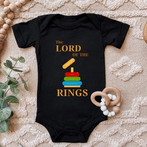 Op de afbeelding: Zwarte baby romper met de tekst "De Heer der Ringen" in gouden letters. Een kleurrijk stapelring speelgoed is afgebeeld onder de tekst.