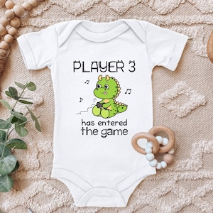 Op de afbeelding: Witte baby romper met een groene dinosaurus die een videogamecontroller vasthoudt. De romper zegt "Player 3 has entered the game".