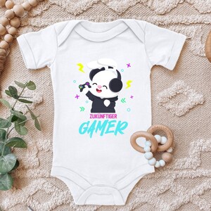 Könnte beinhalten: Weißer Baby Strampler mit einem Panda, der Kopfhörer trägt und einen Videospiel-Controller hält. Der Text "ZUKUNFTIGER GAMER" ist auf dem Strampler gedruckt.