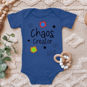 Könnte beinhalten: Ein blauer Baby-Strampler mit dem Text "Chaos Creator" in schwarzer Schrift. Der Strampler hat ein rotes, gelbes und grünes Sonnenmotiv und einen schwarzen Stern.