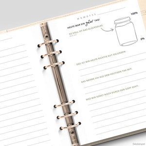 May include: A white planner with a gold binder and a black and white illustration of a jar with a lid. The text "HEUTE WAR EIN guter TAG!" is written in black. The text "SO VOLL IST DAS GL&Uuml;CKSGLAS:  Mole aus!" is written in black. The text "DAS IST MIR HEUTE RICHTIG GUT GELUNGEN:" is written in black. The text "DAS NEHME ICH AUS DEM HEUTIGEN TAG MIT:" is written in black. The text "WAS MIR SONST NOCH DURCH DEN KOPF GEHT:" is written in black. The text "Dekobeispiel" is written in black.