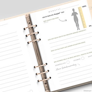 May include: A white planner with a gold binder and a section for daily reflection. The planner includes a scale with a figure and the text "Heute war ein mieser Tag!" (Today was a bad day!).
