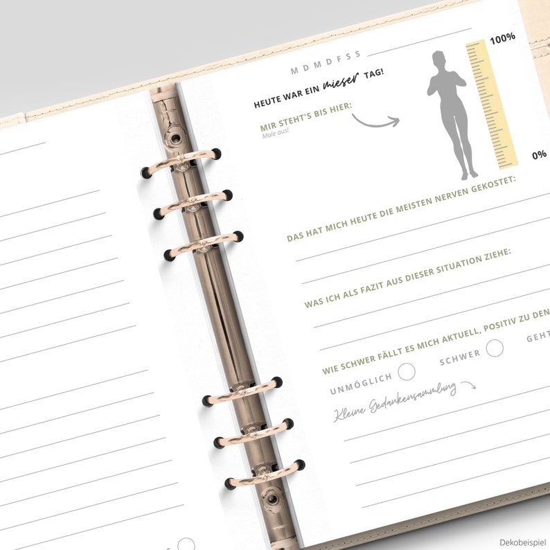 May include: A white planner with a gold binder and a section for daily reflection. The planner includes a scale with a figure and the text "Heute war ein mieser Tag!" (Today was a bad day!).