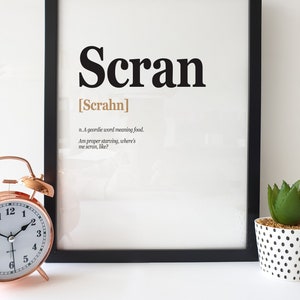 May include: A black framed print with the word "Scran" in bold black letters and the definition "n. A geordie word meaning food. Am proper starving, where's me scran, like?" in smaller black letters. The word "Scran" is written in a larger font size than the definition.