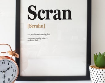 May include: A black framed print with the word "Scran" in bold black letters and the definition "n. A geordie word meaning food. Am proper starving, where's me scran, like?" in smaller black letters. The word "Scran" is written in a larger font size than the definition.