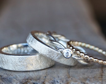 May include: Three silver wedding bands, one with a small diamond, on a wooden surface.