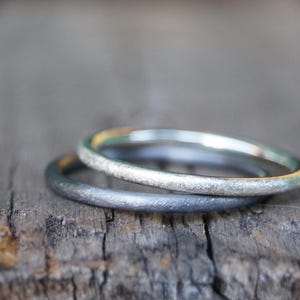 Könnte beinhalten: Zwei Silberringe, einer übereinander gestapelt, liegen auf einer verwitterten Holzoberfläche. Der obere Ring hat eine hellere, reflektierendere Oberfläche, während der untere Ring dunkler und matter erscheint. Die Ringe sind wahrscheinlich Eheringe oder Modeschmuck.