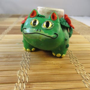 Peut inclure: Figurine verte faite à la main ressemblant à un personnage de dessin animé. La figurine a des pieds jaunes, des accents rouges et un objet cylindrique beige sur le dos. La figurine est posée sur un tapis en bambou.