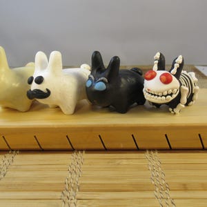 May include: Four whimsical bunny figurines in various colors, including cream, white, and black, with unique facial features. One has a mustache, another has skeleton detailing, and another has red eyes. They are displayed on a wooden board.
