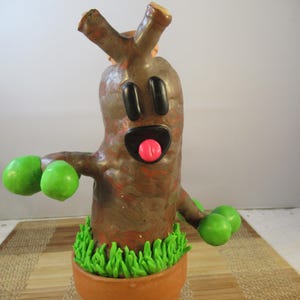 May include: A whimsical, handmade tree figurine with a cartoonish design. The brown tree has black eyes, a pink tongue, and green spheres for hands. It sits in a terracotta pot with green grass.