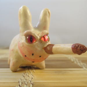 May include: A whimsical ceramic bunny figurine with red eyes and a cigarette. The bunny is off-white with pink accents on the cheeks and mouth. The cigarette is brown and white. The figurine is on a wooden surface.