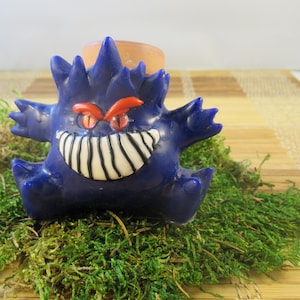 Puede incluir: Figura de dibujos animados azul oscuro con una sonrisa amenazante y ojos rojos. La figura está posada sobre un lecho de musgo verde. La figura tiene una cabeza puntiaguda y brazos extendidos. Un pequeño cilindro marrón está detrás de la figura.