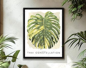 May include: A framed art print featuring a large, green and yellow variegated Monstera leaf. The print is titled "Thai Constellation" in a minimalist font. The artwork is displayed against a neutral background, with surrounding greenery.