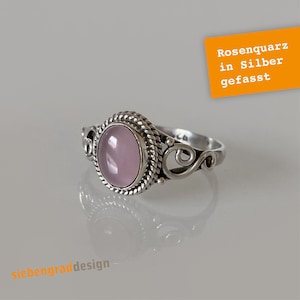 Könnte beinhalten: Ein silberner Ring mit einem rosafarbenen Rosenquarz-Stein, der in eine gedrehte Silberband gefasst ist. Der Ring liegt auf einer weißen Oberfläche. "Rosenquarz in Silber gefasst" steht in orange auf einem weißen Rechteck.
