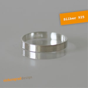 Könnte beinhalten: Ein silberner Ring mit einem flachen, rechteckigen Band. Der Ring ist hochreflektierend, mit dem Text "Silber 925" in einem orangefarbenen Rechteck. Der Ring wird auf einer reflektierenden Oberfläche dargestellt.