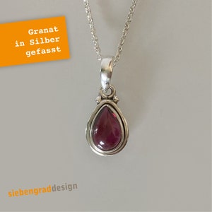 Könnte beinhalten: Eine silberne Halskette mit einem tropfenförmigen Granat-Anhänger. Der Anhänger ist in einer silbernen Fassung gefasst und hat eine zarte Kette.