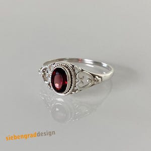 Könnte beinhalten: Ein Silberring mit einem oval geschliffenen, tiefroten Granatstein in einer Perlenfassung. Die Ringschiene zeigt auf beiden Seiten des Mittelsteins komplizierte, durchbrochene Schnörkelmuster. Der Text "siebengradesign" ist in der unteren Ecke zu sehen.
