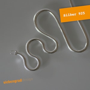 Könnte beinhalten: Eine silberne Schlangenkettenkette aus 925er Sterlingsilber. Die Kette liegt auf einer grauen Oberfläche. Der Text "Silber 925" ist auf einem gelben Etikett sichtbar.