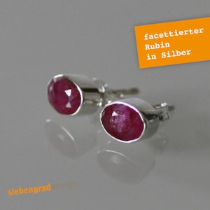 Könnte beinhalten: Ein Paar silberne Ohrstecker mit ovalen, facettierten Rubin-Edelsteinen. Die Rubine sind tiefrot und in eine polierte Silberfassung gefasst. Auf der Abbildung steht "facetierter Rubin in Silber".