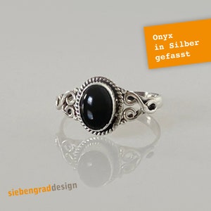 Könnte beinhalten: Ein silberner Ring mit einem schwarzen Onyxstein, der in einem filigranen, verzierten Design gefasst ist. Der Ring liegt auf einer weißen Oberfläche mit dem Text "Onyx in Silber gefasst" in einem gelben Kasten.