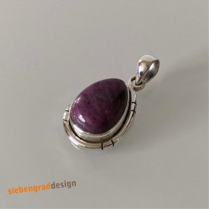 Könnte beinhalten: Ein silberner Anhänger mit einem großen, ovalen, tiefvioletten Edelstein. Der Edelstein ist in einer silbernen Lünette mit geätzten Linien gefasst. Der Anhänger hat eine Silberschlaufe für eine Kette. Der Text "siebengraddesign" befindet sich unten links.