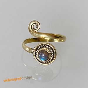 Könnte beinhalten: Ein goldener Ring mit einem geschwungenen Band und einem runden, schillernden Edelstein, der in einer goldenen Fassung sitzt. Die Fassung ist mit einem seilartigen Muster verziert.